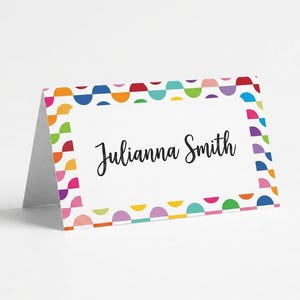 May include: A folded white place card with the name "Julianna Smith" in black script. The card is bordered with a colorful pattern of semi-circles in red, orange, yellow, green, blue, purple, and pink.
