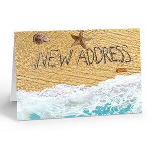 May include: A folded greeting card featuring a sandy beach scene with the text NEW ADDRESS written in the sand. The design includes a brown starfish, a spiral seashell, and a small orange shell, with white ocean waves washing onto the shore.