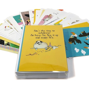 May include: A collection of greeting cards with various illustrations and text. The cards are displayed in a clear plastic case. One card features a cartoon animal with the text "Now's the time to celebrate! So have fun, live it up and enjoy life."