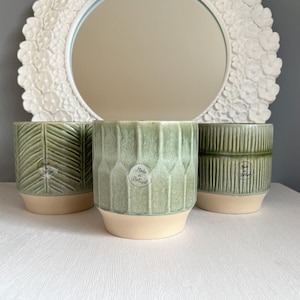 May include: Three green ceramic plant pots with textured designs, including leaf and geometric patterns, sit on a white surface. Each pot has a beige base and a label that reads "Made in Portugal". A white floral mirror is in the background.