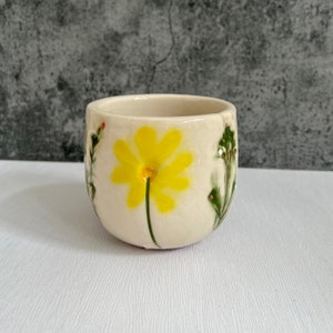 May include: A small, off-white ceramic cup with a hand-painted yellow daisy and green foliage. The cup has a rounded shape and is decorated with delicate floral accents in red and green. The cup is suitable for holding small beverages.