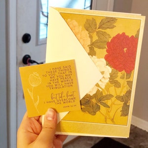 May include: A greeting card with a floral design featuring red, white, and yellow flowers. The card is held with a smaller yellow card with text that reads "I have overcome the world." The background is a door and window.