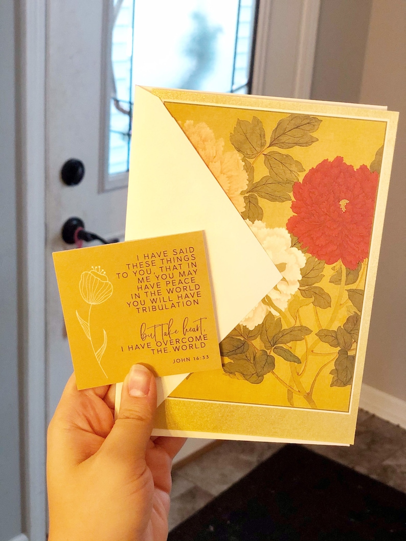 May include: A greeting card with a floral design featuring red, white, and yellow flowers. The card is held with a smaller yellow card with text that reads "I have overcome the world." The background is a door and window.