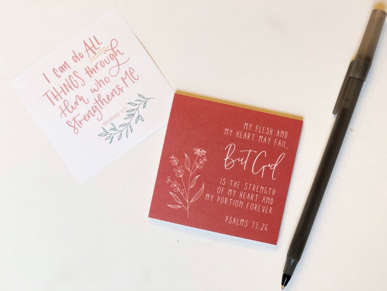 May include: Two square cards with inspirational quotes and floral illustrations. One card is white with pink text that reads "I can do all things through Him who strengthens me." The other card is red with white text that reads "My flesh and my heart may fail, but God..." A pen is also in the image.