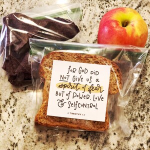May include: A packed lunch featuring a red apple, a slice of bread with a printed message, and a bag of dark-colored snacks. The message on the bread reads "For God did not give us a spirit of fear but of power, love & self-control 2 Timothy 1:7."