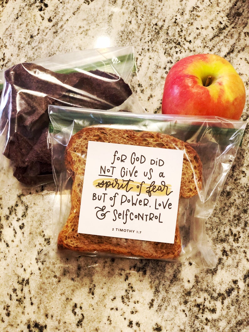 May include: A packed lunch featuring a red apple, a slice of bread with a printed message, and a bag of dark-colored snacks. The message on the bread reads "For God did not give us a spirit of fear but of power, love & self-control 2 Timothy 1:7."