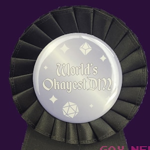 May include: A black ribbon with a purple button that says "World's Okayest DM" in white text. The button has a white dice and stars on it. The ribbon says "Gay Nerd Goods" in pink text.