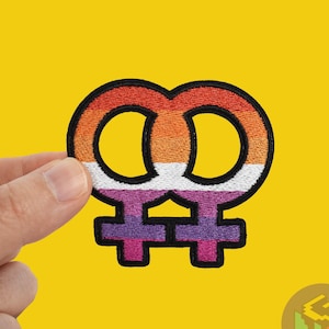 Op de afbeelding: Een close-up van een lesbische pride-patch met twee in elkaar grijpende vrouwelijke symbolen in oranje, roze, wit en paars.