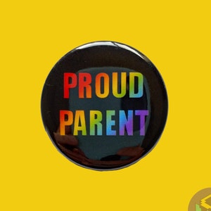 Puede incluir: Un botón redondo y negro con las palabras "PROUD PARENT" en texto con los colores del arcoíris. El botón está sobre un fondo amarillo brillante. Un pequeño logotipo está en la esquina inferior derecha.