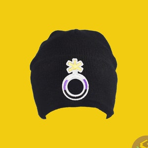 Op de afbeelding: Zwarte beanie met een geborduurd genderneutraal symbool in paars en geel op de voorkant.