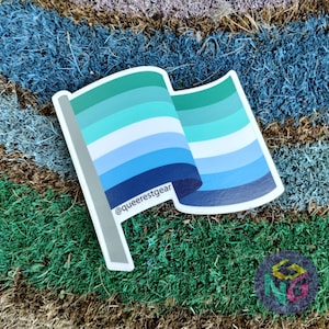 May include: A blue, green, and white pride flag sticker with the text "@queerestgear" on it. The sticker is on a green and brown surface.