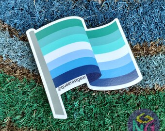 May include: A blue, green, and white pride flag sticker with the text "@queerestgear" on it. The sticker is on a green and brown surface.