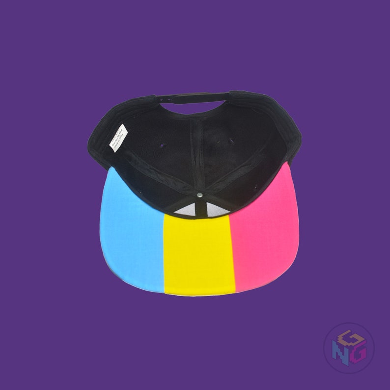 Black flat bill snapback hat. The brim has the pansexual pride flag on both sides and the front of the hat has the word “PAN” in pink, yellow, and blue letters. Underside view