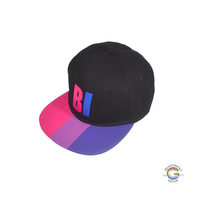 Black flat bill snapback hat. The brim has the bisexual pride flag on both sides and the front of the hat has the word “BI” in pink and blue. Front left view