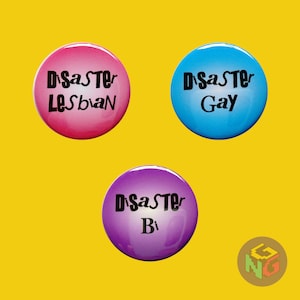 Pode incluir: Três botões redondos com a palavra "Disaster" em texto preto sobre um fundo colorido. Os botões são rosa, azul e roxo. O botão rosa diz "Disaster Lesbian", o botão azul diz "Disaster Gay" e o botão roxo diz "Disaster Bi".
