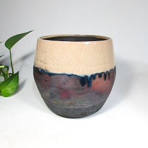 May include: A handcrafted ceramic tumbler with a unique design. The upper portion is a cream color with a cracked texture, transitioning to a deep blue and reddish-brown midsection, and a dark gray base. The tumbler has a wide, rounded shape.