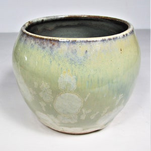 May include: A handcrafted ceramic bowl with a rounded shape and a wide opening. The bowl features a light green glaze with subtle blue and yellow hues, and a dark brown rim. White crystalline patterns adorn the exterior.
