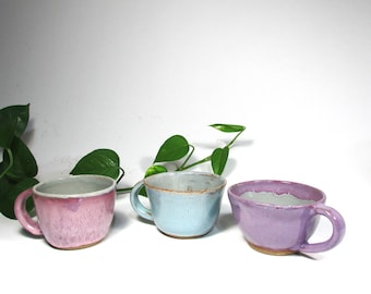 May include: Three ceramic mugs in different colors: pink, light blue, and purple. Each mug has a unique glaze and a slightly different shape. The mugs are arranged in a row on a white surface.