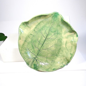May include: A ceramic leaf-shaped dish with a detailed leaf vein pattern. The dish is primarily light green with darker green accents, creating a naturalistic aesthetic. The edges are gently curved.