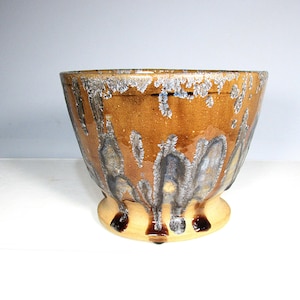 May include: A handcrafted ceramic bowl with a wide, open top and a rounded base. The bowl features a warm brown glaze with unique dripping patterns in shades of gray and white, creating a textured appearance. The base is a lighter tan color.