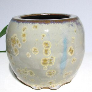May include: A ceramic vase with a rounded shape and a wide opening. The vase is primarily a pale gray color, with yellow and brown crystalline patterns. The rim is a darker brown, and the base has a textured edge.