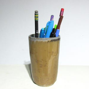 May include: A ceramic pencil holder in a warm, earthy brown tone. The holder contains various pens and pencils, including a black pencil with green and yellow stripes, a blue pen, and a red-topped pencil. The holder has a slightly textured surface.