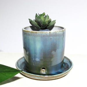 May include: A ceramic planter with a blue and green glaze, holding a green succulent. The cylindrical pot sits on a matching saucer with a drainage hole. The planter is a decorative item for indoor plants.