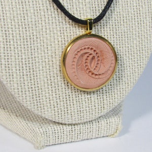 May include: A round pendant necklace with a gold-colored frame and a pinkish-tan center. The center features an embossed spiral design. The necklace hangs on a black cord, displayed on a neutral-colored bust.