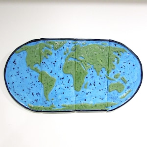 May include: A ceramic world map with a blue ocean and green continents. The map is divided into several rectangular sections and has a dark blue border. The ocean is speckled with dark blue dots.