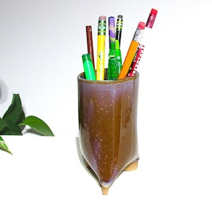 May include: A brown ceramic pencil holder with a purple glaze, holding various pencils and pens. The holder has a unique, triangular base with three small feet. The pencils and pens are in various colors, including yellow, green, and red.