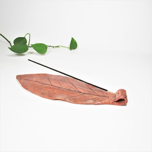 May include: A ceramic incense holder shaped like a leaf, in shades of reddish-brown. The leaf-shaped holder has detailed vein patterns and a small, rolled-up end. Two black incense sticks rest on the holder, and green leaves are in the background.