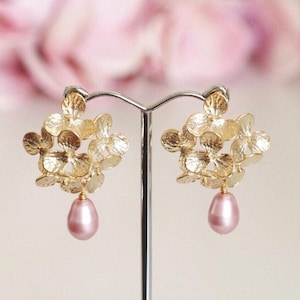 Peut inclure: Boucles d'oreilles dorées avec un motif floral en grappe et une perle rose pâle en forme de goutte. Les boucles d'oreilles sont présentées sur un support argenté, sur un fond rose flou.