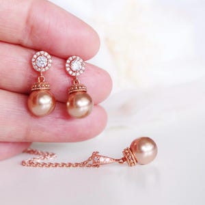 May include: A rose gold jewellery set comprising earrings and a necklace. The earrings have a halo of small clear stones around a central clear stone, with a large, round, peach-coloured pearl. The necklace features a similar pearl pendant.