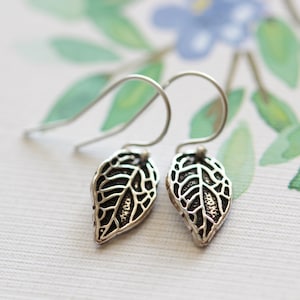 Puede incluir: Un par de pendientes de plata con colgantes en forma de hoja. Las hojas tienen un diseño delicado e intrincado.