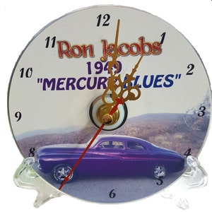 May include: A white clock face with black numbers and gold hands. The clock face has a picture of a purple 1949 Mercury car. The text "Ron Jacobs 1949 "MERCURABLUES" is printed on the clock face.