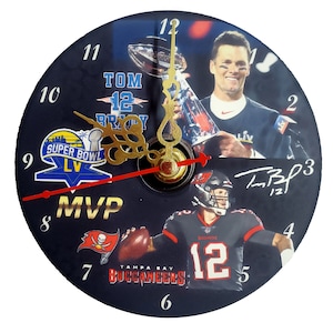 May include: A black and white clock with a picture of Tom Brady holding a trophy. The clock has a black background with white numbers and a red second hand. The clock also has a picture of Tom Brady in a Tampa Bay Buccaneers uniform. The clock has the words "Super Bowl LV" and "MVP" on it.