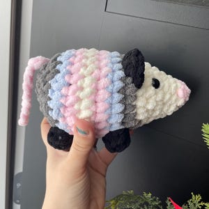 May include: A plush, crocheted opossum toy featuring a white face, black ears, and black feet. The body is crafted with thick, textured yarn in alternating vertical stripes of grey, light blue, white, and pink, with a long, thin pink tail.