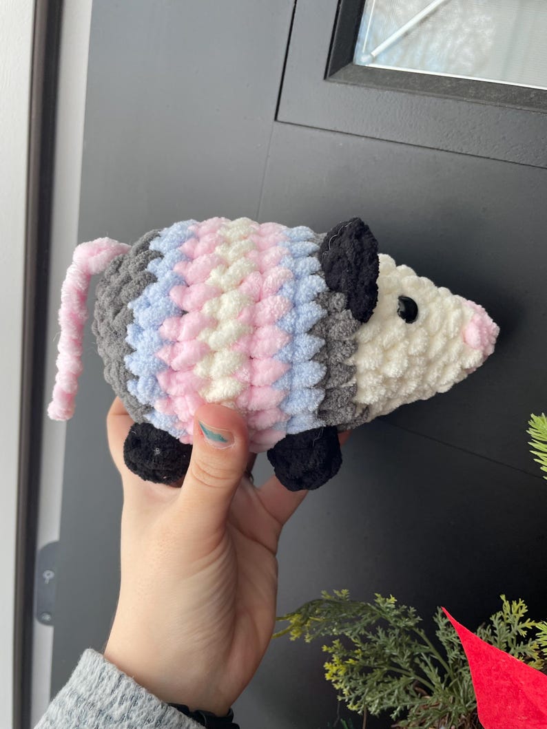 May include: A plush, crocheted opossum toy featuring a white face, black ears, and black feet. The body is crafted with thick, textured yarn in alternating vertical stripes of grey, light blue, white, and pink, with a long, thin pink tail.