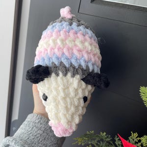 May include: A plush, crocheted opossum toy featuring a white face, a pink nose, and black ears. The body is crafted from soft, textured yarn in horizontal stripes of grey, light blue, white, and pink, topped with a small pink tuft of yarn.