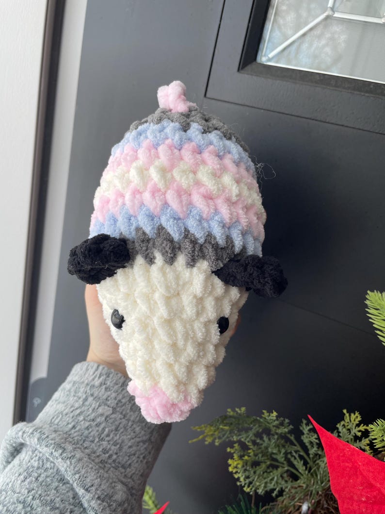 May include: A plush, crocheted opossum toy featuring a white face, a pink nose, and black ears. The body is crafted from soft, textured yarn in horizontal stripes of grey, light blue, white, and pink, topped with a small pink tuft of yarn.