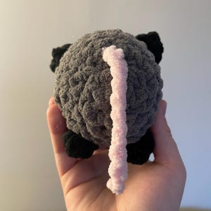 May include: A small, round, crocheted plush toy shaped like an animal. The body is made of textured dark grey yarn, featuring small black crocheted feet and ears, and a long, thin, light pink crocheted tail extending from the centre of the back.