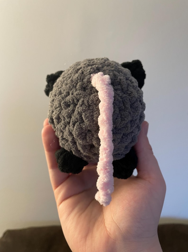 May include: A small, round, crocheted plush toy shaped like an animal. The body is made of textured dark grey yarn, featuring small black crocheted feet and ears, and a long, thin, light pink crocheted tail extending from the centre of the back.