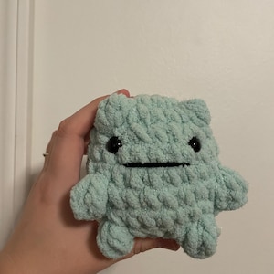 May include: A light blue, handmade crochet creature with black button eyes and a stitched mouth. The plush toy has a textured, chunky appearance and small arms and legs. The background is a plain white wall.