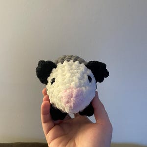May include: A small, handmade crochet plush toy shaped like an opossum. The creature features a white face, a soft pink nose, black ears, and black feet, with a textured grey body. It has two small, round, dark plastic eyes set into the white yarn.
