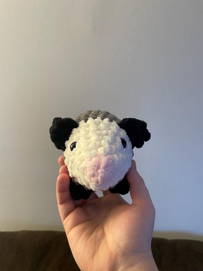May include: A small, handmade crochet plush toy shaped like an opossum. The creature features a white face, a soft pink nose, black ears, and black feet, with a textured grey body. It has two small, round, dark plastic eyes set into the white yarn.