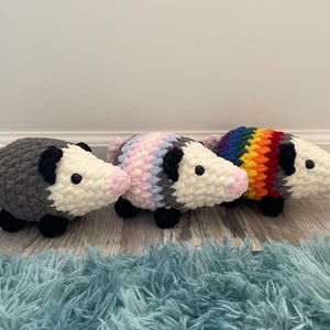 May include: Three crocheted plush opossums arranged in a row. The first is dark grey, the second features pastel pink, blue, and white stripes, and the third displays a vibrant rainbow pattern. Each has a white face, black ears, black feet, and a pink nose.