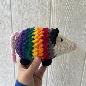 May include: A crocheted plush opossum featuring a white head with a pink nose and black ears. The body is crafted with horizontal stripes of purple, blue, green, yellow, orange, and red yarn. It has a light pink tail and small black feet.