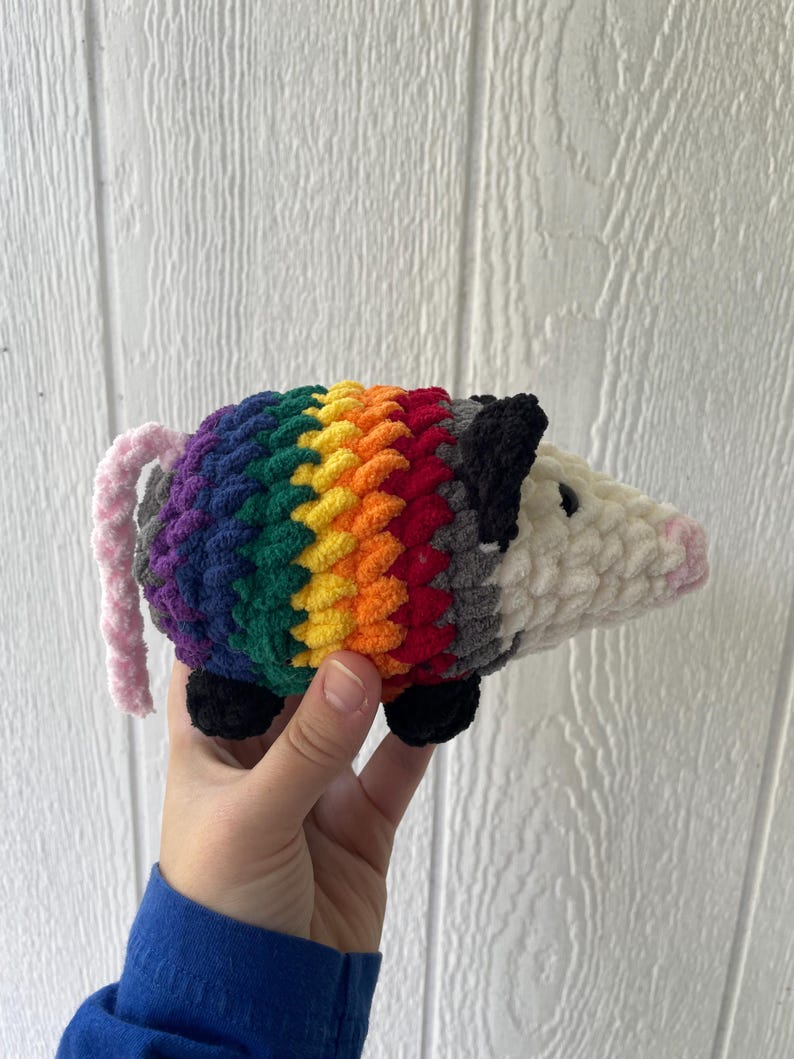 May include: A crocheted plush opossum featuring a white head with a pink nose and black ears. The body is crafted with horizontal stripes of purple, blue, green, yellow, orange, and red yarn. It has a light pink tail and small black feet.