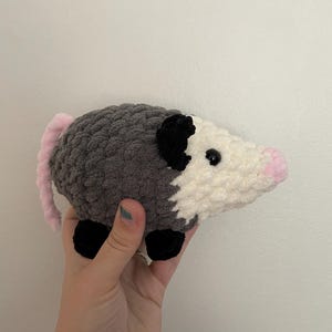 May include: A handmade crochet plush toy shaped like an opossum. The body is crafted from dark grey yarn with a white face, black ears, and black feet. It features a small pink nose and a long, light pink tail extending from the back of the body.
