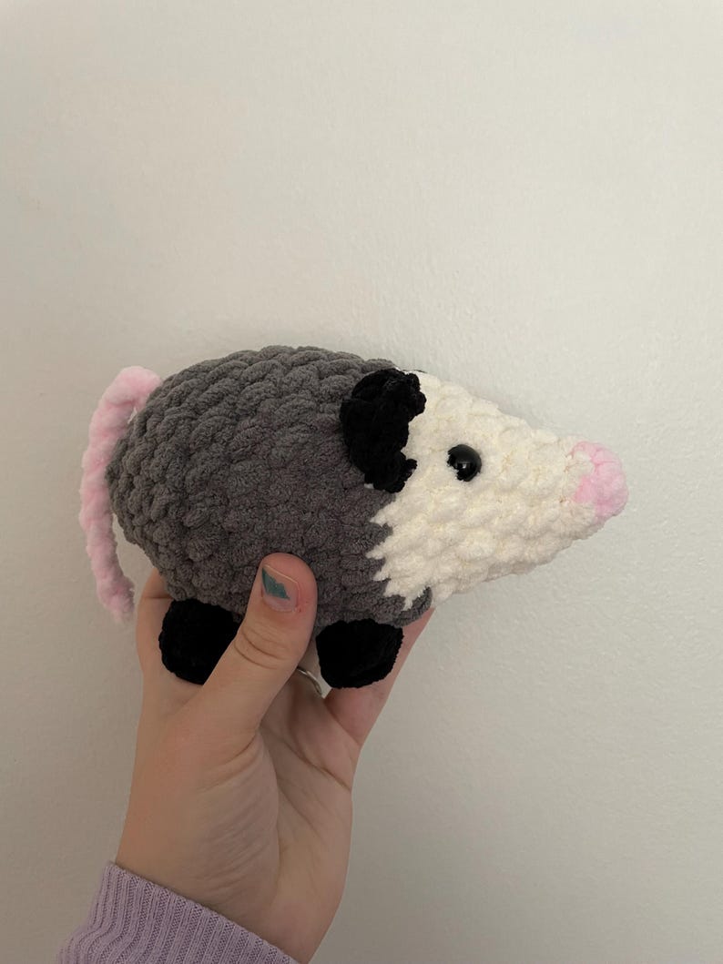 May include: A handmade crochet plush toy shaped like an opossum. The body is crafted from dark grey yarn with a white face, black ears, and black feet. It features a small pink nose and a long, light pink tail extending from the back of the body.